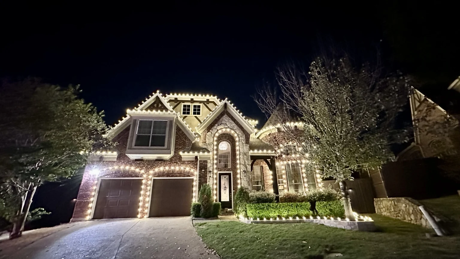 Large brick and stone home outlined with warm white Christmas lights across the roofline, garage, entry and landscaping